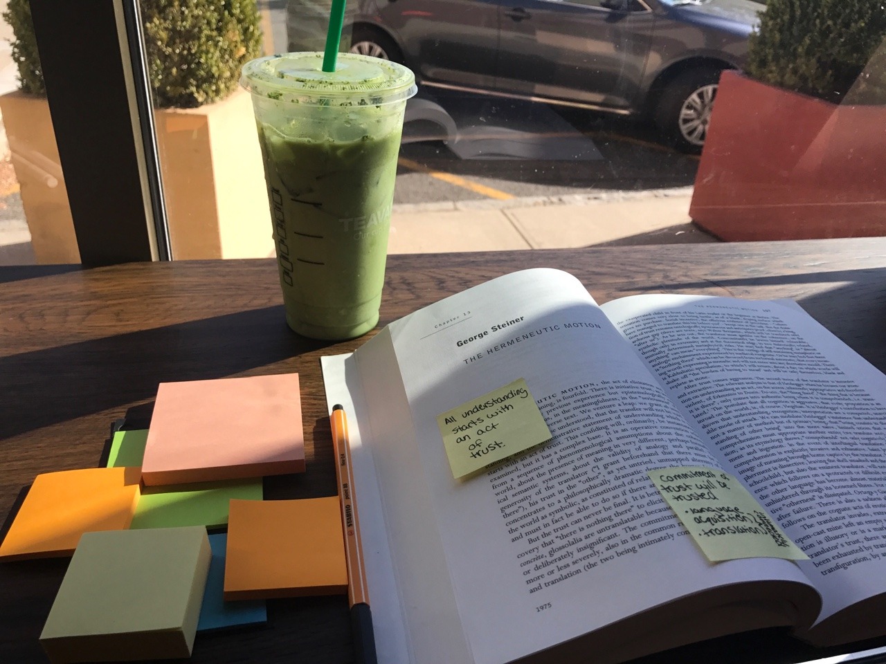 A translation theory textbook is open in front of a window. Next to it is an iced matcha latte and a pile of post-it notes.
