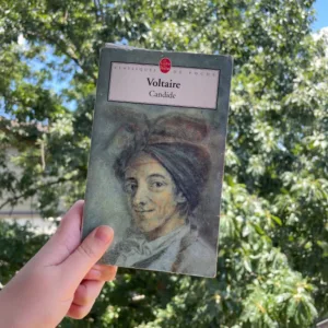 A paperback copy of Cadide by Voltaire in front of a tree on a sunny day.